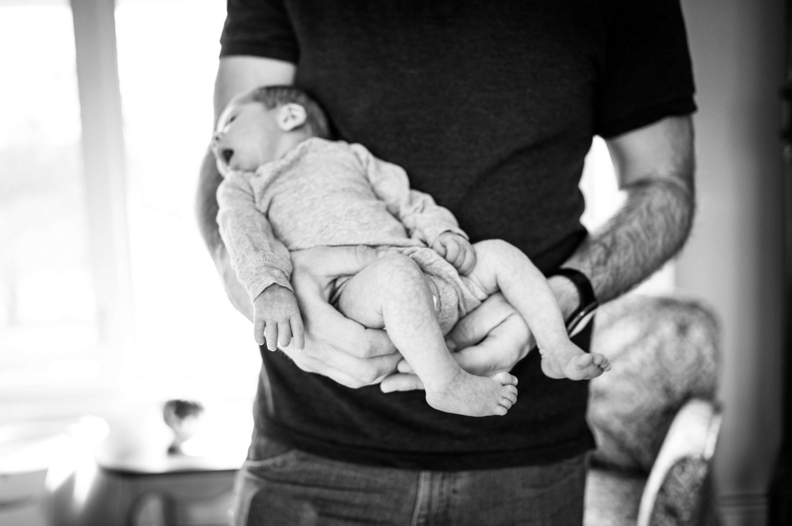 A newborn baby lays in dad's arms in a black and white image in a onesie before visiting pediatric dentists in new haven