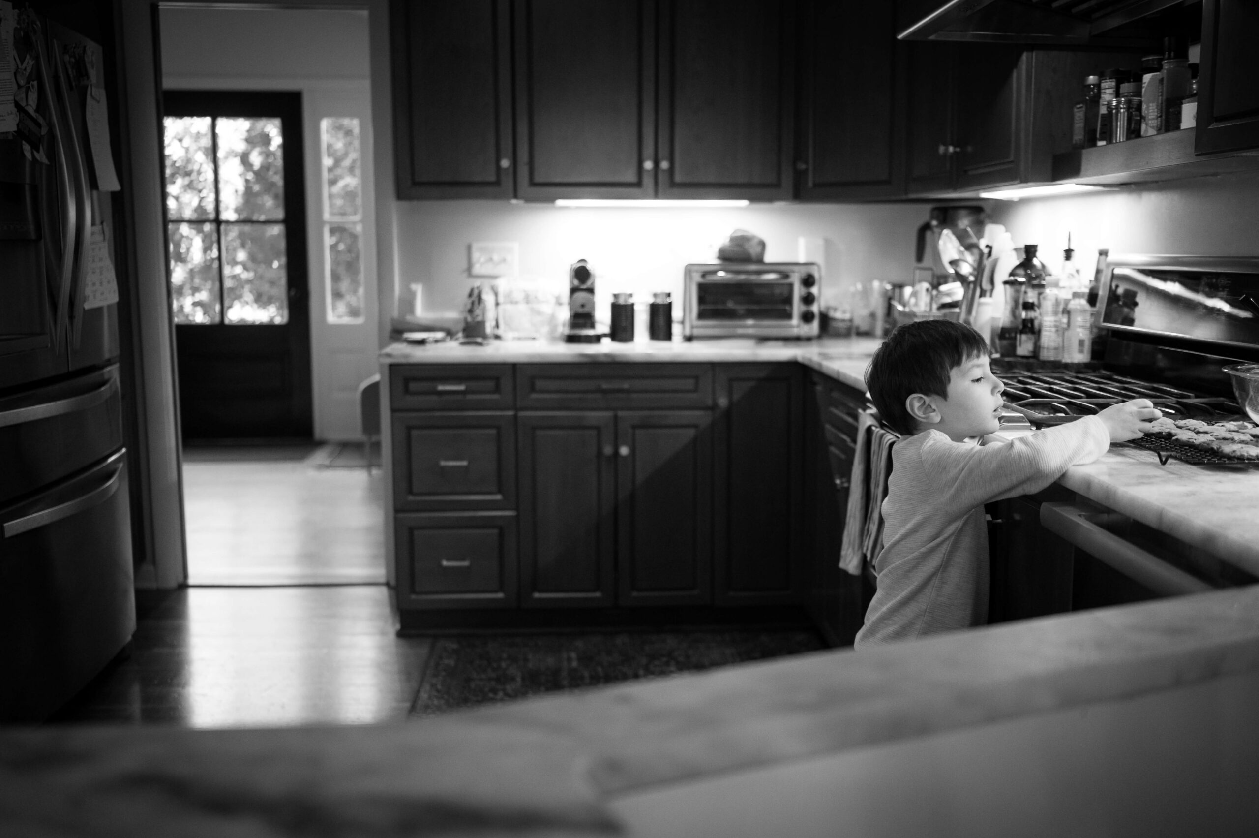 A toddler boy reaches up to the kitchen counter for cookies after visiting Grace and Haven in black and white