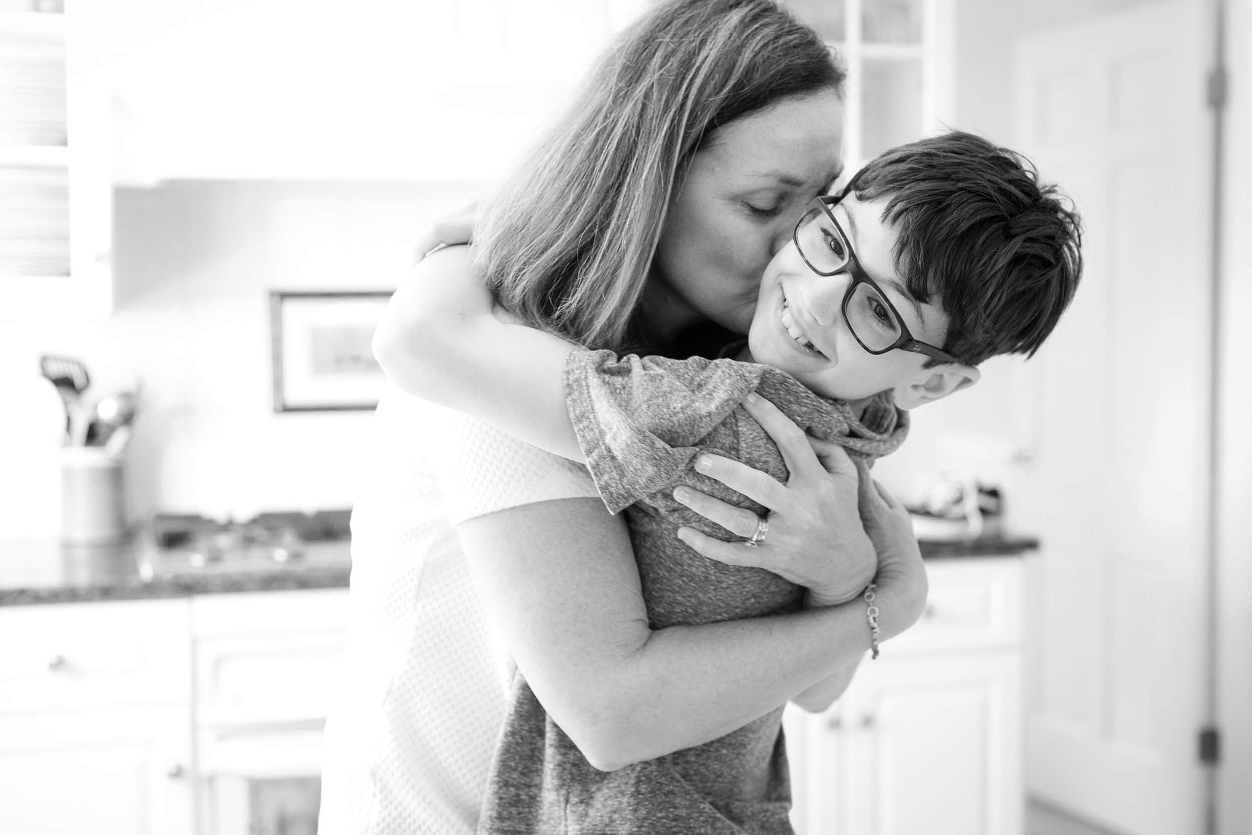 A smiling young boy with glasses is hugged and kissed in a kitchen by mom in black and white