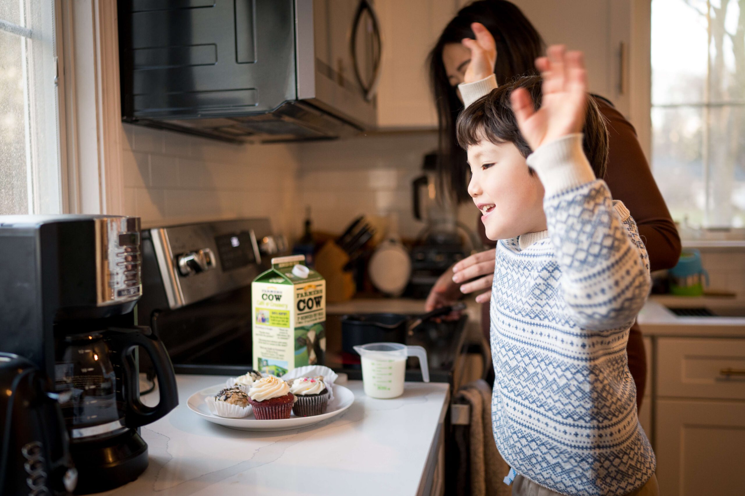 A happy toddler boy in a blue sweater celebrates over a plate of baked goods in a kitchen with mom