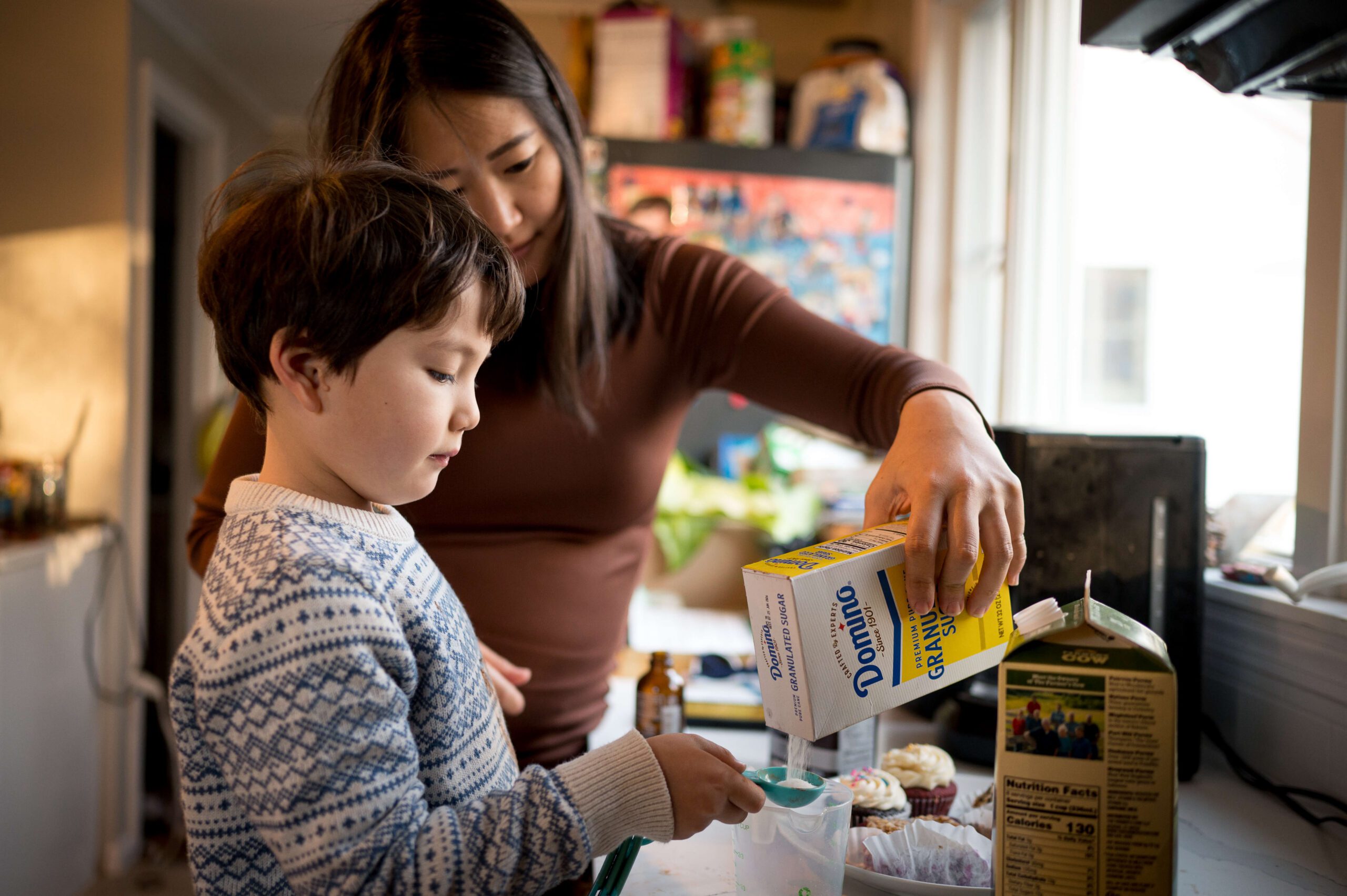 A mom helps a toddler boy in a blue sweater measure sugar in the kitchen after some mommy and me classes in new haven ct