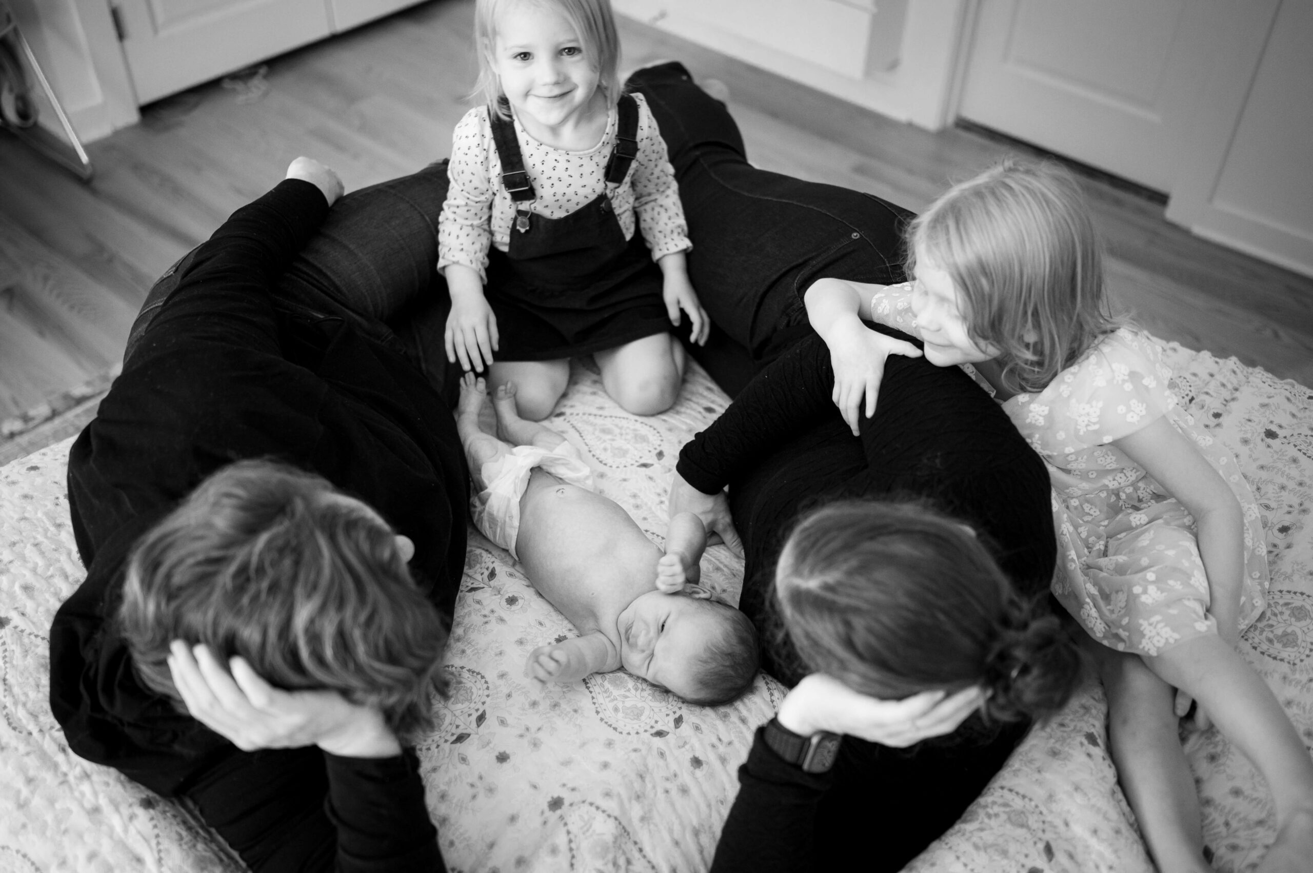 Moms and two toddler girls lay on a bed on top of each other around an awake newborn
