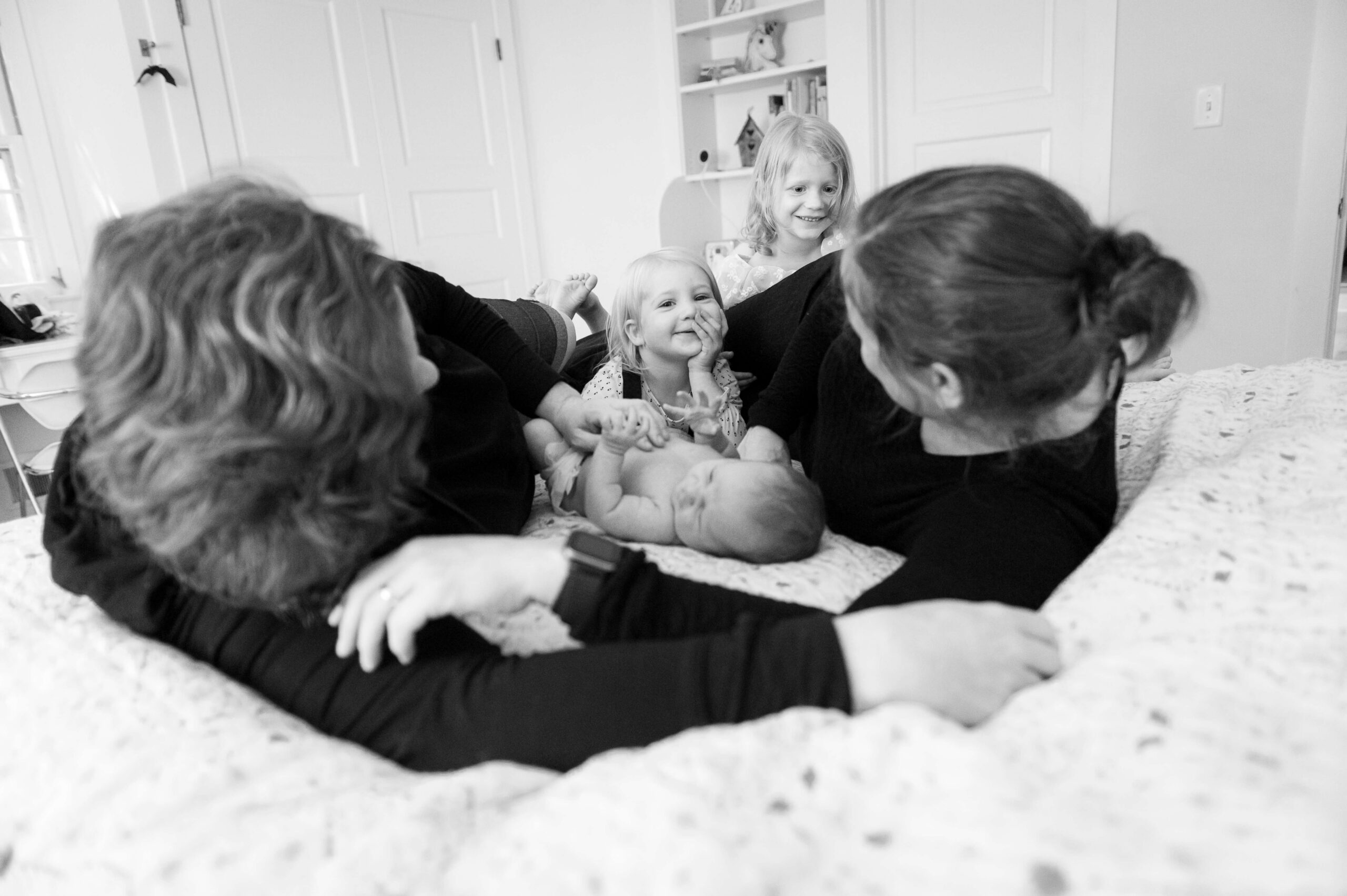 Happy toddler girls lay across a bed with their moms and newborn sibling between them after visiting montessori schools in new haven county
