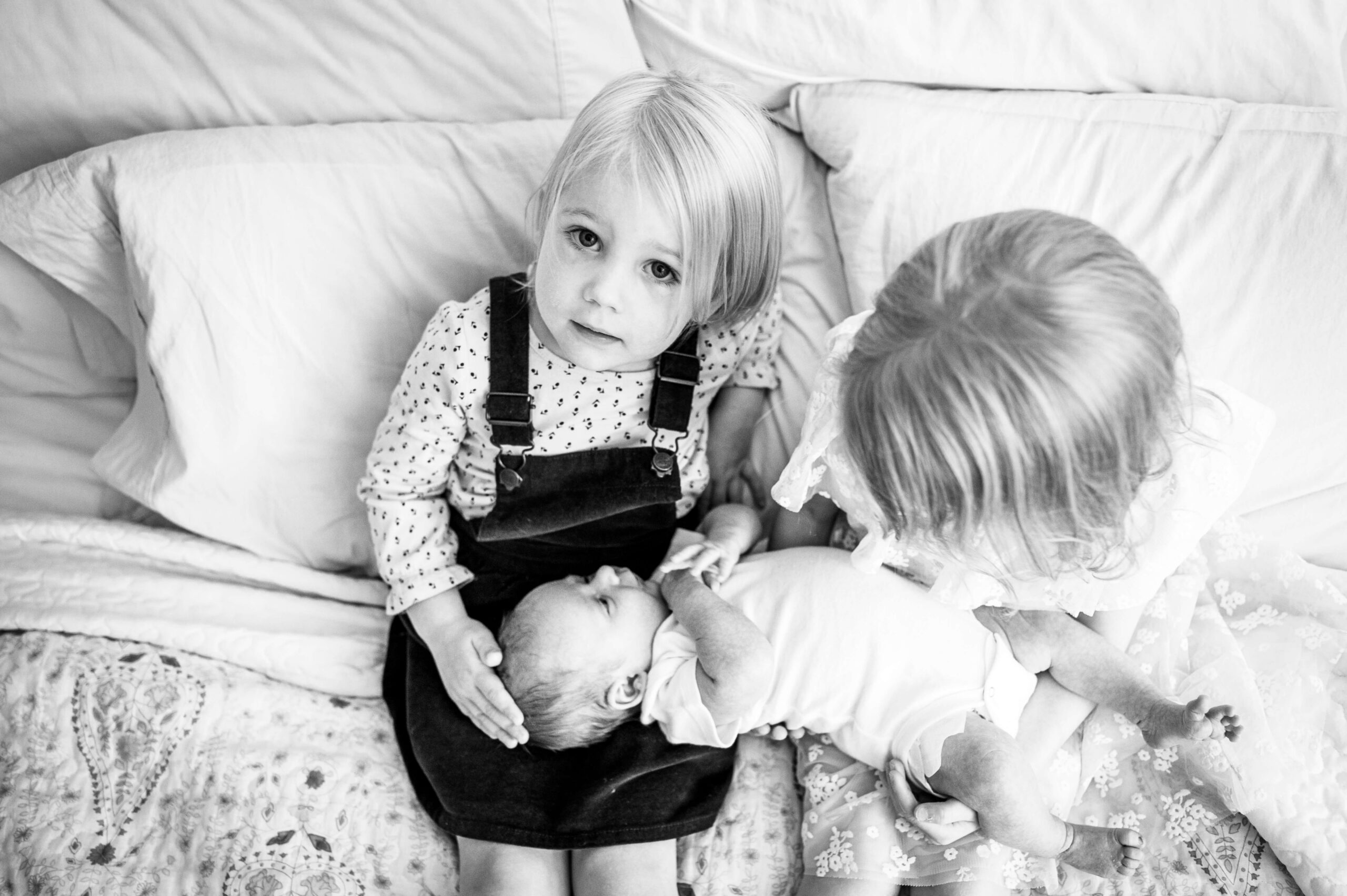 Toddler sisters sit on a bed with their newborn sibling in their laps in black and white after visiting montessori schools in new haven county