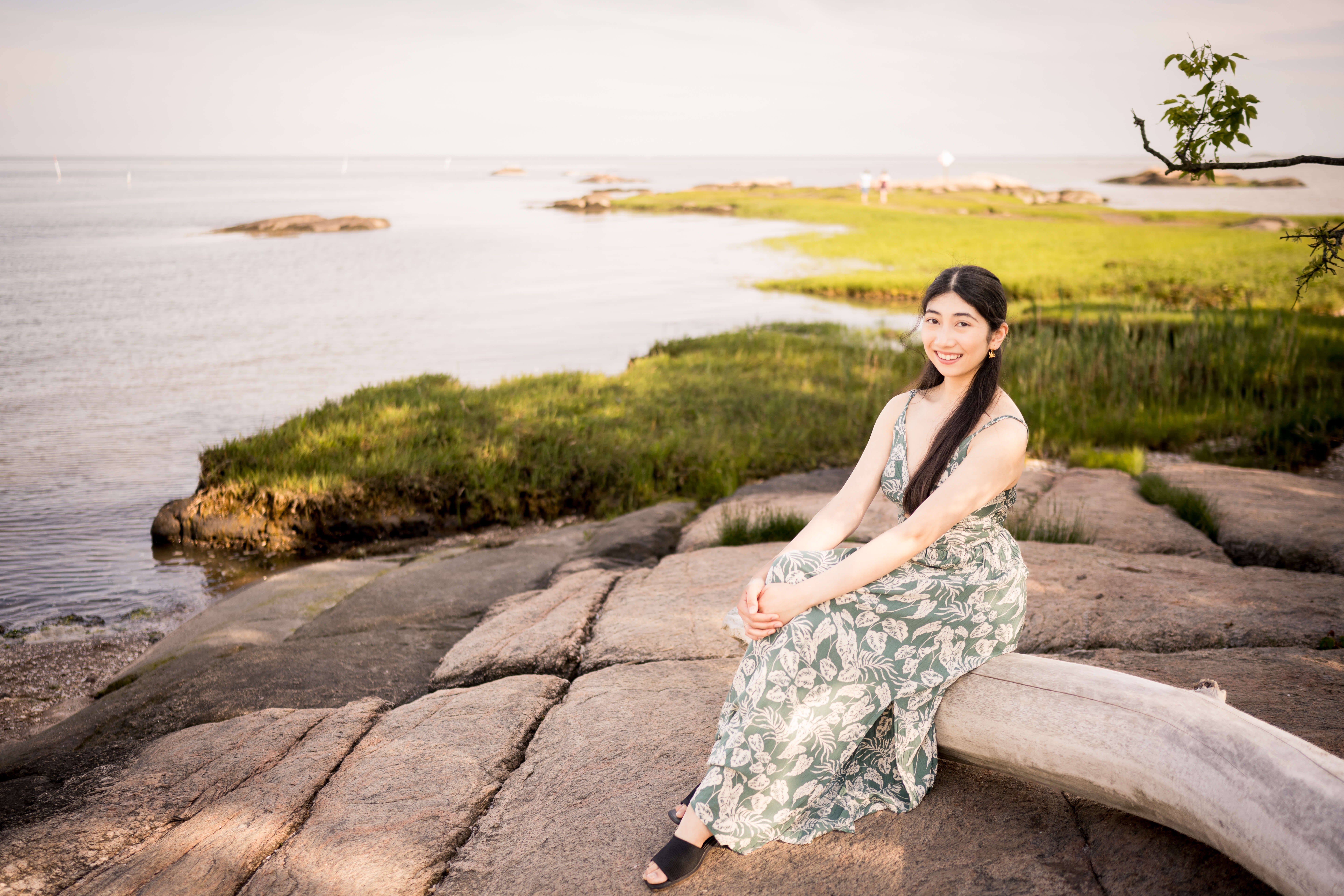 A happy high school senior in a floral print dress sits on driftwood by the water smiling with hands on her knee