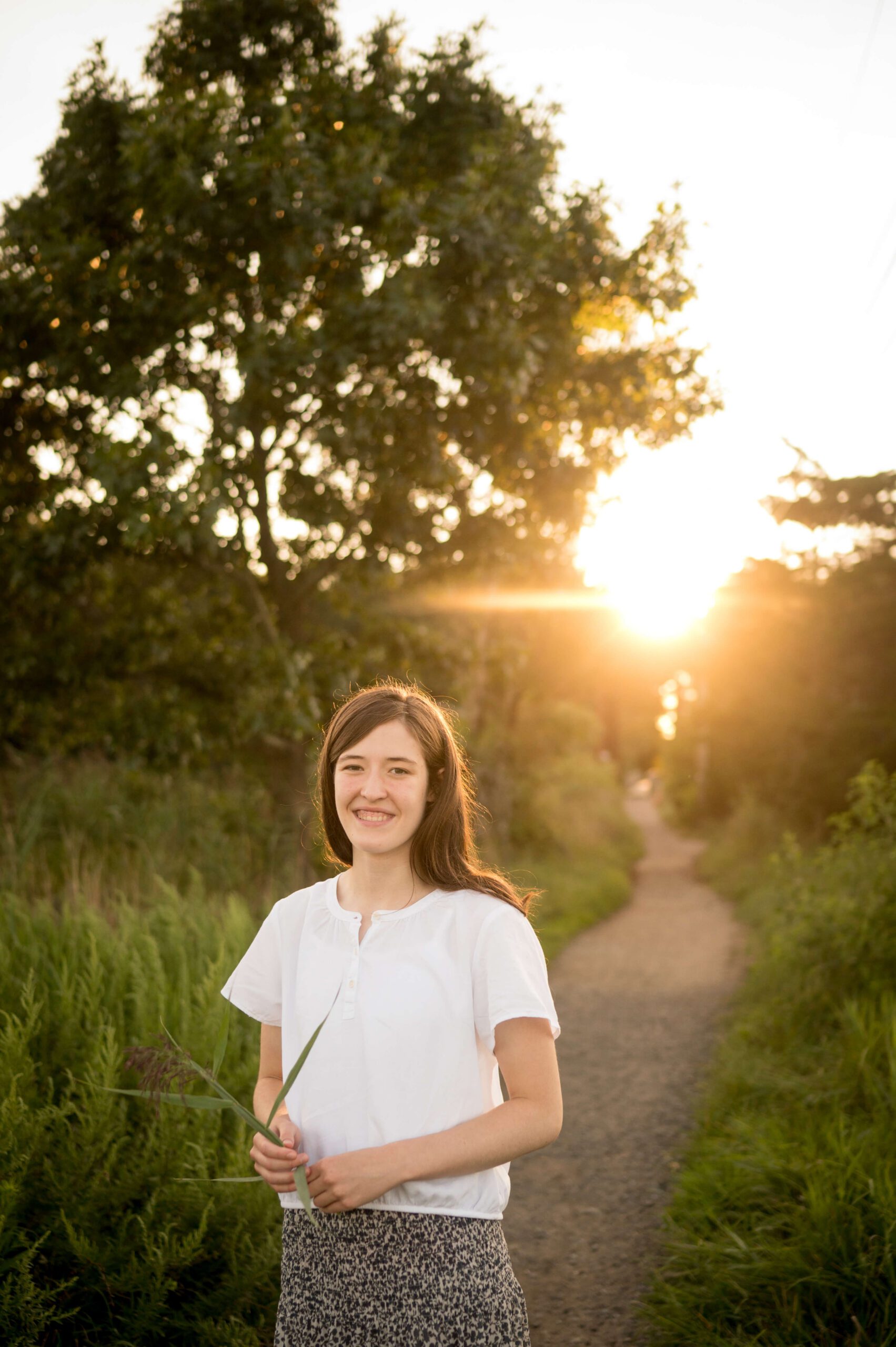 A smiling high school senior in a white blouse wlaks a park trail at sunset before exploring party venues in new haven ct