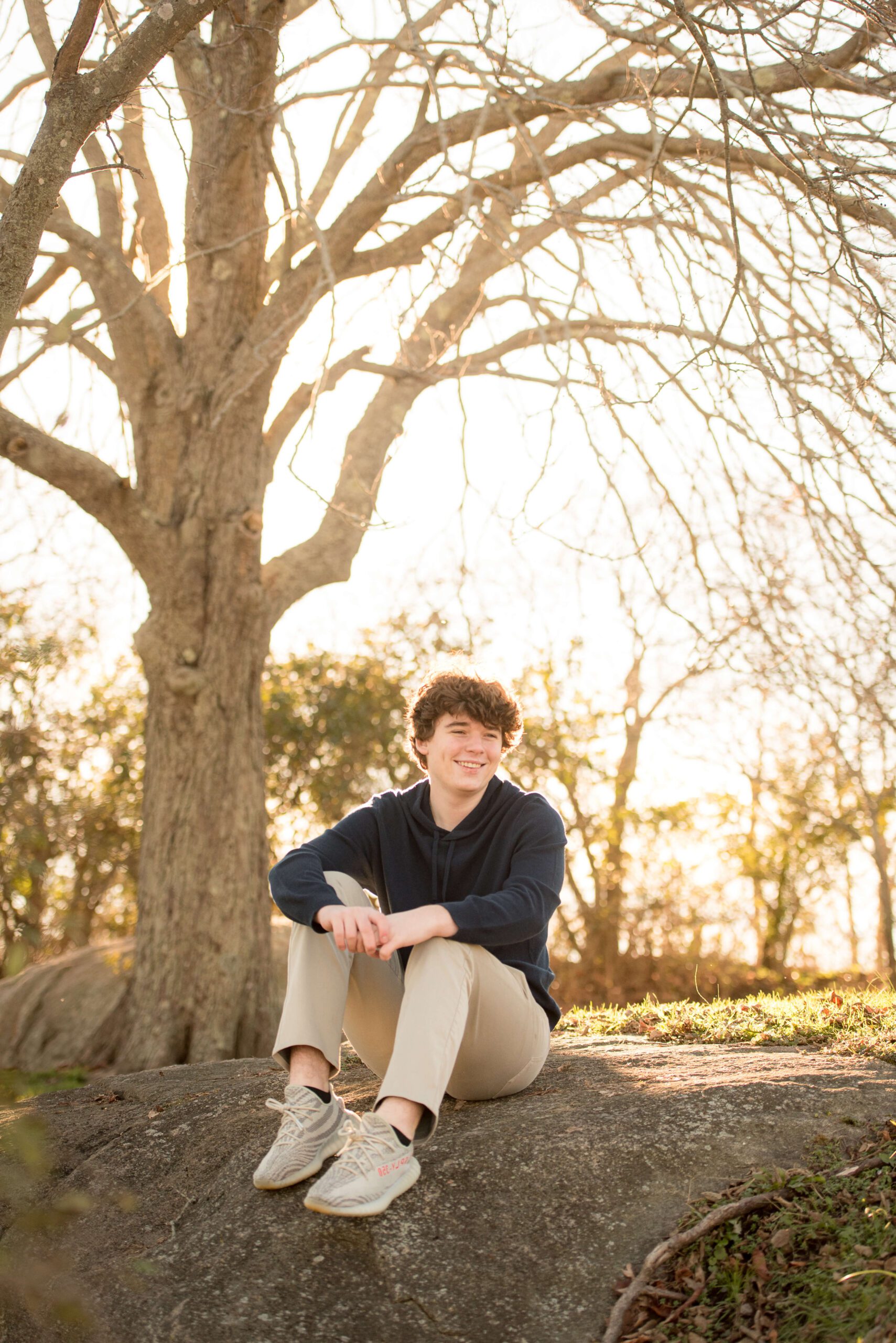 A high school senior smiles while sitting on a boulder in a park at sunset in a blue hoodie before visiting party venues in new haven ct