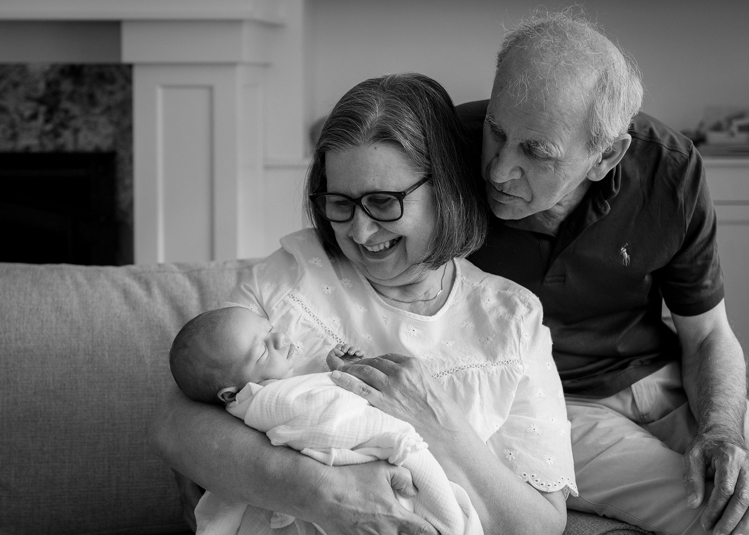 A smiling grandma cradles her sleeping newborn grandchild while sitting on a couch as grandpa looks over her shoulder from the arm rest