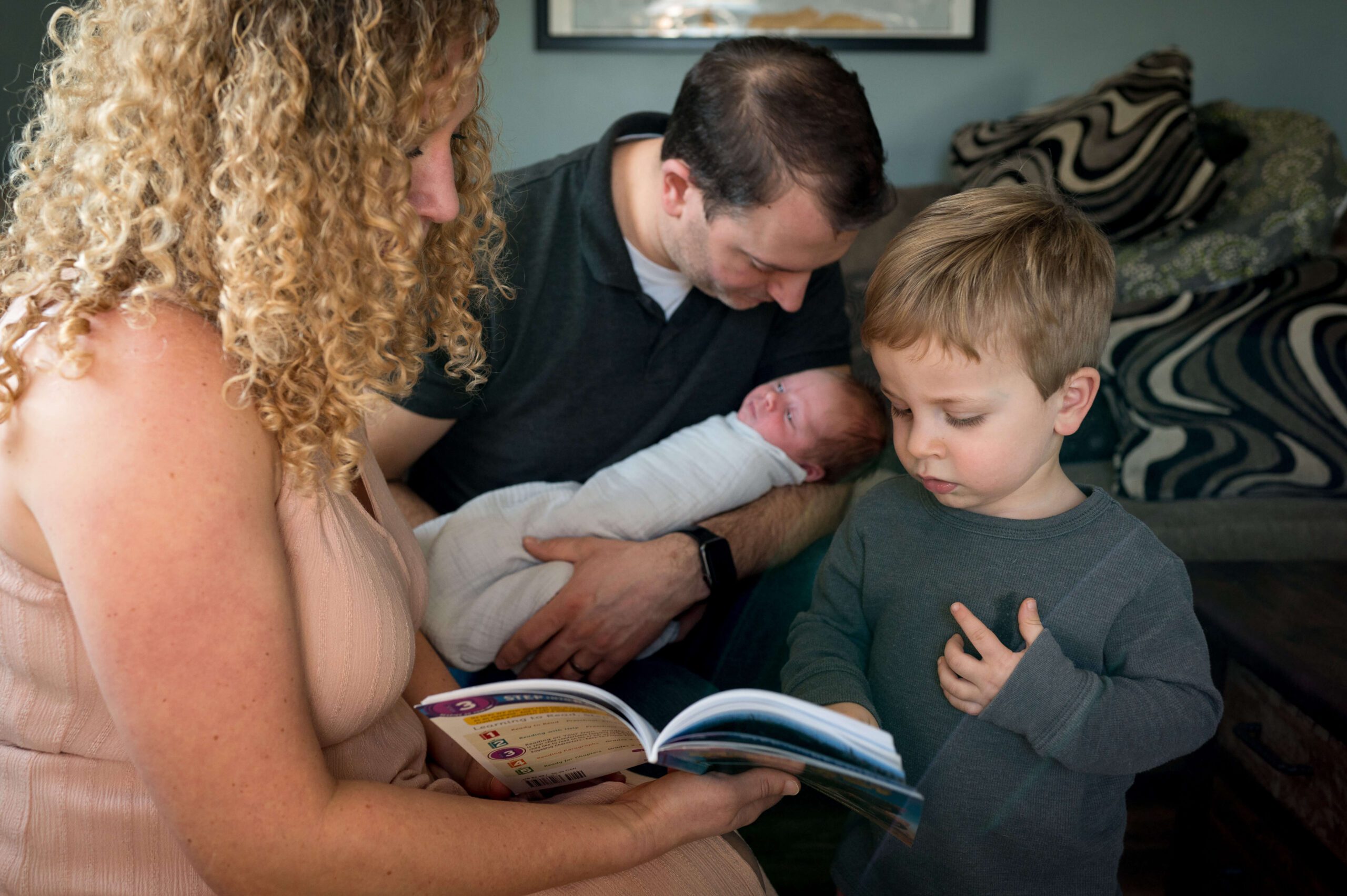 A young boy reads a book with mom on a couch as dad cradles a newborn baby behind them after visiting preschools in new haven