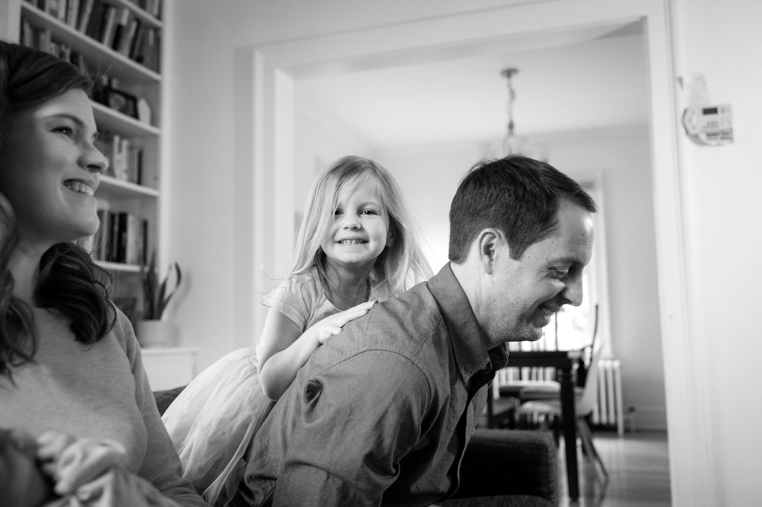A happy toddler girl climbs on dad's back as he laughs on a couch